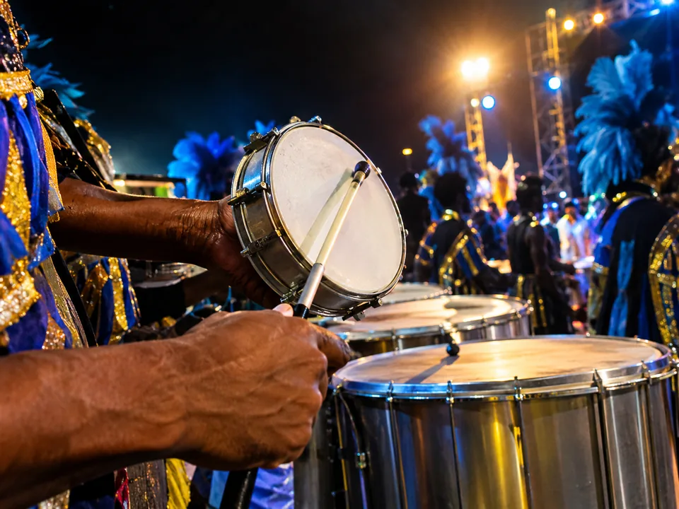 Hands striking a small tamborim drum in front of a row of silver surdo drums